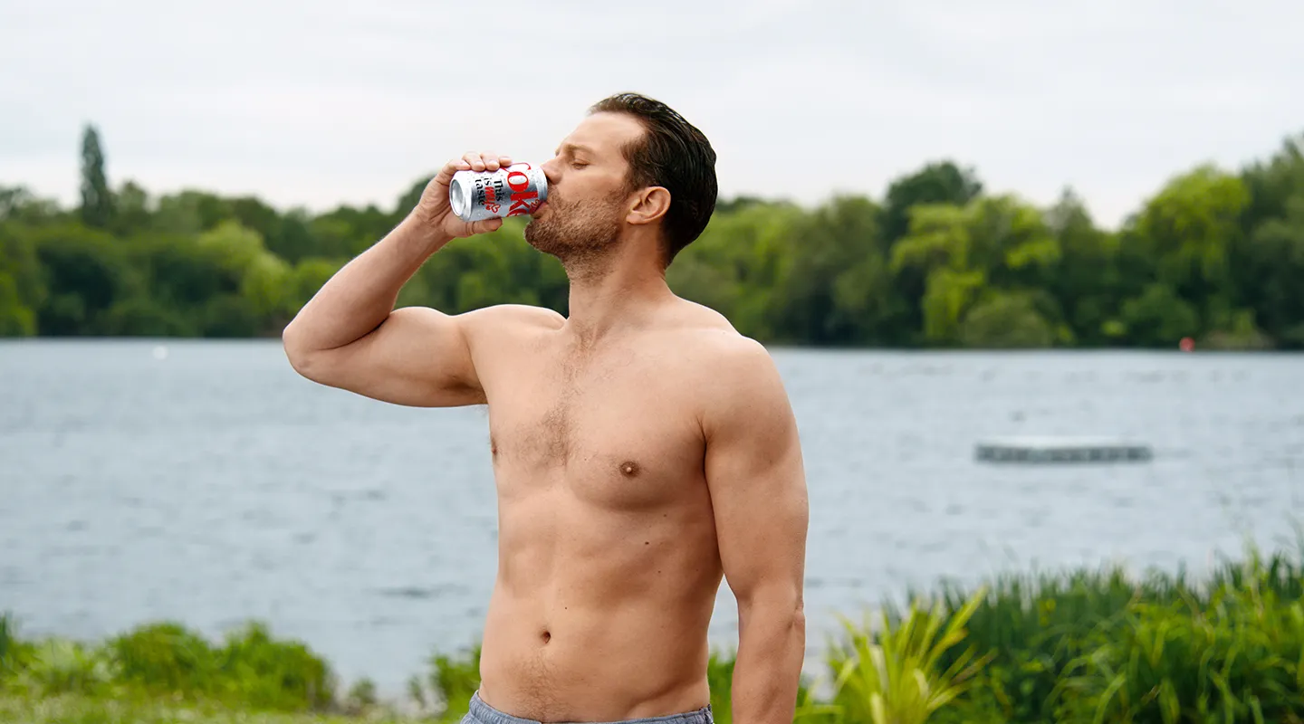 A shirtless man drinks a can of Diet Coke beside a lake.